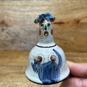 May include: A whimsical ceramic bell figurine with a surprised expression. The bell features a white base with blue and brown painted details, including a cat and a face with orange cheeks and blue hair. The bell is held in a hand.