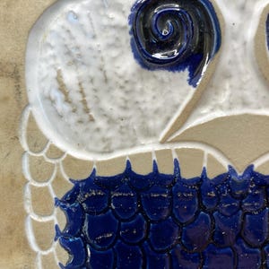 May include: Close-up of a ceramic owl design. The owl features a white head with a dark blue spiral eye and a blue scale-like pattern on the lower portion. The background is a light tan color.
