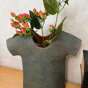 May include: A dark gray ceramic vase shaped like a t-shirt with short sleeves and a rounded neckline. The interior of the vase is a reddish-brown color. A sprig of green stems with small, round, orange and red berries is placed inside the vase.