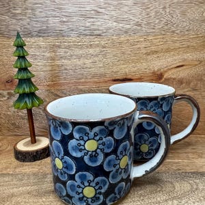 May include: Two ceramic mugs featuring a dark blue background with a repeating pattern of light blue flowers with yellow centers, complemented by a small, carved wooden evergreen tree figurine with a brown trunk and a green conical top.