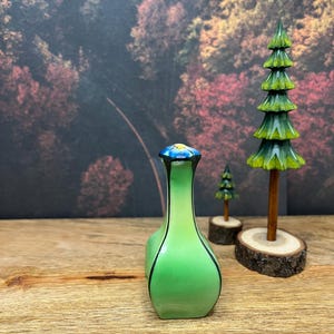 May include: A green ceramic vase with a blue and yellow floral design on the stopper. The vase has black trim and is set on a wooden surface. Two wooden decorative trees are in the background.