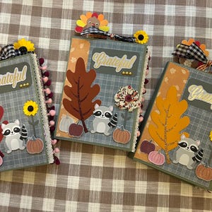 May include: Three handmade journals with autumn-themed decorations. Each journal features a plaid pattern, a large leaf, a raccoon, pumpkins, and the word "Grateful." They are adorned with sunflowers and turkey accents.