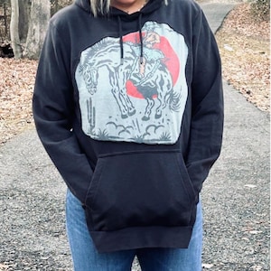 May include: Black hoodie with a kangaroo pocket and a drawstring hood. The front features a graphic of a cowboy riding a horse, set against a red circle and a light blue background. The design includes a cactus. The person is wearing blue jeans.