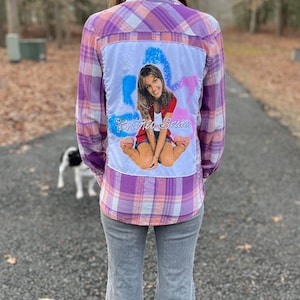 May include: A plaid button-down shirt in shades of purple, pink, and white, with a custom graphic on the back. The graphic features a person and the text "Britney Spears". The person is wearing gray jeans.