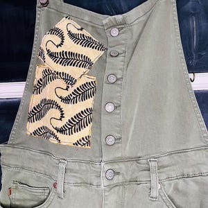May include: Olive green denim overalls with a bib pocket featuring a yellow and black fern pattern. The overalls have adjustable straps, metal buttons, and a front pocket. The overall design is casual and stylish.