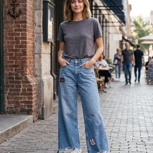 May include: A charcoal gray crew neck t-shirt paired with light blue wide-leg denim jeans featuring a raw, frayed hem. The jeans include a small rectangular patch on the upper right thigh and a smaller patch on the lower left leg, worn with white sneakers.