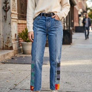 May include: A cream-colored long-sleeve sweater paired with blue denim jeans featuring vertical seams and colorful patchwork panels at the lower legs. The jeans are secured with a black leather belt, and the outfit is completed with off-white sneakers.