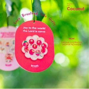 May include: A red oval-shaped air freshener with the text "Joy to the world the Lord is come" and a floral design with cartoon figures. The fragrance type is Coconut. The words "Love Breathed into Gifts" are written on the top.