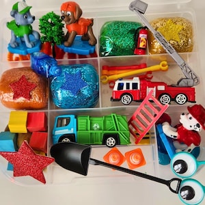 May include: A clear plastic container filled with various toys and craft supplies. Includes toy trucks, fire truck, toy figures, glittery play dough, and a shovel. The toys are in a variety of colors, including red, green, blue, and yellow.