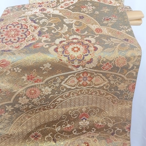 May include: Close-up of a decorative textile with an intricate floral pattern. The fabric features a blend of gold, brown, and muted green hues, with accents of red and purple. The design includes stylized flowers and geometric shapes, creating a rich, textured appearance.