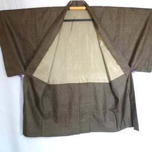 以下が含まれることがあります： 小さなチェック柄の茶色の着物。着物は開いており、ベージュの裏地が見えます。袖は広く、衣類は木製のハンガーに吊るされています。着物は柔らかい生地で作られています。