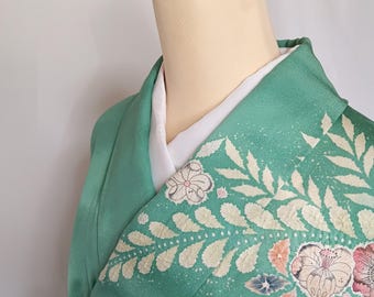 May include: A teal kimono with floral and leaf patterns in white, pink, and orange. The garment is displayed on a mannequin, with a white underlayer visible at the neckline. A red and gold clasp secures the kimono. The text "A302ZT" is visible.