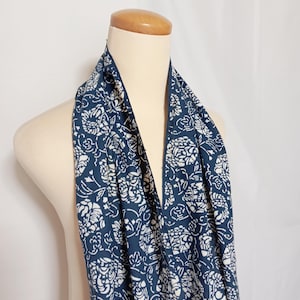 May include: A navy blue scarf with a white floral design is draped over a mannequin. The scarf displays a pattern of white flowers and leaves against a dark blue backdrop. The fabric looks soft and lightweight, suitable for various styles.