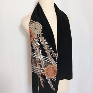 May include: Black scarf with an ornate floral and leaf design in shades of orange, white, and brown. The scarf is draped over a mannequin, showcasing the detailed embroidery and elegant style. The scarf is a fashion accessory.