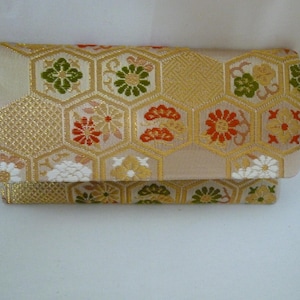 Op de afbeelding: Een beige clutch met een gouden geometrisch patroon en bloemaccenten in rood, groen en wit. De clutch heeft een gevouwen ontwerp en een textuur, geschikt voor het dragen van kleine items.