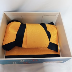 May include: A folded yellow and black fabric garment with a subtle geometric star pattern, neatly packed inside a rectangular white cardboard box. The box features a blue decorative border with Japanese text printed on the front side panel.