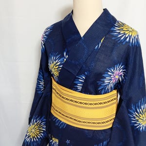 Puede incluir: Un yukata azul oscuro con un patrón repetitivo de motivos florales en forma de estrella amarillos y azules. La prenda se ajusta a la cintura con un fajín obi amarillo ancho y texturizado que incluye finas rayas horizontales negras y patrones geométricos.