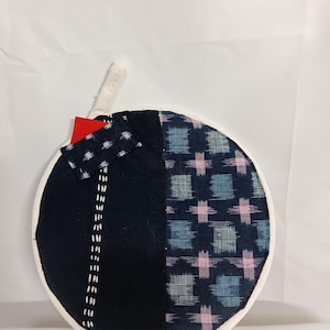 以下が含まれることがあります： 白い縁取りの丸いキルティング鍋つかみ。デザインは、白いステッチが施された黒い生地のパッチワークと、模様のある青とピンクの正方形が特徴です。小さな赤いタグと白いループが取り付けられています。