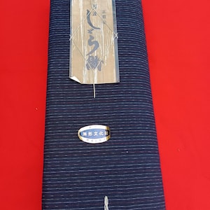 Könnte beinhalten: Ein gefaltetes Stück dunkelblauen Stoffes mit dünnen weißen und roten horizontalen Streifen. Ein Papieranhänger mit japanischem Text ist mit einer Schnur befestigt. Ein kleiner ovaler Aufkleber in Blau und Weiß ist ebenfalls angebracht.