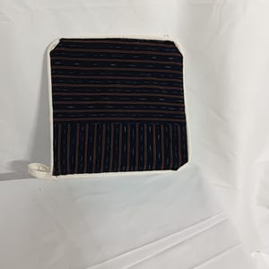 Puede incluir: Un guante de cocina cuadrado, azul marino y marrón estampado, con borde blanco y una pequeña presilla para colgar. El guante presenta un diseño a rayas con líneas horizontales y verticales.