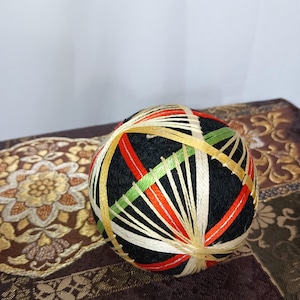Puede incluir: Una bola Temari negra con hilos coloridos en rojo, verde, dorado y blanco. La bola tiene diseños geométricos intrincados. El fondo presenta una tela estampada.