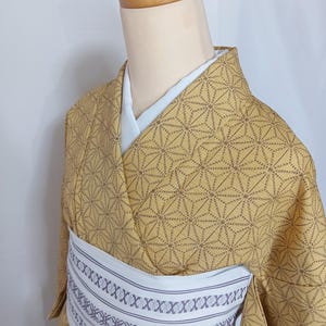 May include: A tan kimono with a geometric pattern and a white obi belt. The kimono is displayed on a mannequin. The obi belt has a repeating pattern of intersecting lines and small decorative elements. The kimono's pattern is made of small, dark brown lines.