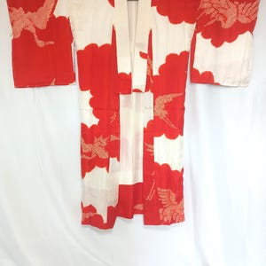 May include: A red and white kimono with a crane pattern. The kimono features a vibrant red background with white cloud-like shapes. The crane design is visible in the sleeves and on the front. The kimono is hanging on a wooden hanger.