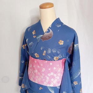 May include: A blue kimono featuring a pattern of small gold and silver flowers and curved shapes, paired with a wide, textured pink obi sash decorated with a white floral print. The garment is displayed on a mannequin torso against a plain background.