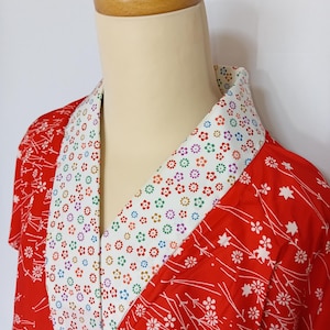 May include: A red kimono featuring a white floral design and a white collar decorated with colourful flowers. The garment is presented on a mannequin, emphasizing its design. The kimono's vibrant colours and intricate details are highlighted.