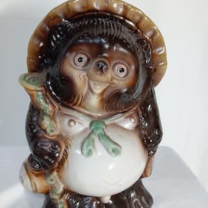 以下が含まれることがあります： 日本の伝統的なタヌキの置物。茶色と白の体、大きな腹、麦わら帽子が特徴です。杖を持ち、緑のリボンをしています。ベースには「A302ZT」の文字があります。