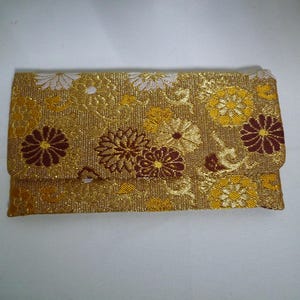Op de afbeelding: Een rechthoekige, goudkleurige clutch met een bloemenpatroon. Het ontwerp toont gouden, witte en kastanjebruine bloemen en bladeren. De clutch heeft een overslagsluiting en is waarschijnlijk gemaakt van een textuurstof.