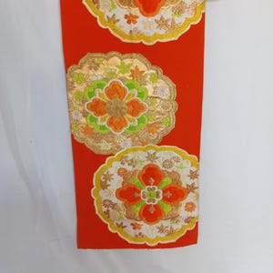 以下が含まれることがあります： 鮮やかなオレンジ色の布地に、3つの刺繍された花のメダリオンがあります。メダリオンは、金、緑、オレンジ色の糸で複雑に刺繍されており、白い背景があります。布は赤いクリップが付いた木製のハンガーから吊り下げられています。長さは約100cmです。