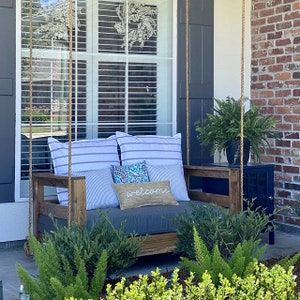 May include: A wooden porch swing with gray cushions and throw pillows. The swing is hanging from the ceiling with rope and has a welcome sign on one of the pillows. The swing is in front of a window with white blinds and a brick wall.