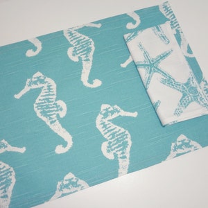 May include: A turquoise blue placemat with a white seahorse design. A white napkin with a starfish design is placed on top of the placemat.
