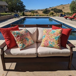 May include: A brown wooden outdoor sofa with white cushions and colorful patterned throw pillows. The sofa is positioned in front of a swimming pool with a view of rolling hills in the background.