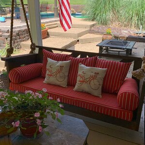 May include: A wooden porch swing with red and white striped cushions and pillows. Two square pillows feature a bicycle design. Two bolster pillows are also red and white striped. The swing is hanging on ropes.
