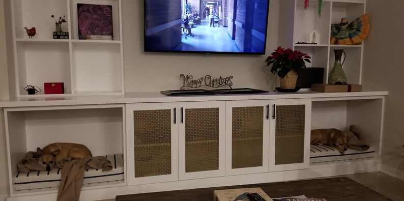 May include: White cabinet with two dog beds, each with a dog sleeping on it. The cabinet has four doors with a woven pattern and black handles. Above the cabinet is a flat screen TV and a white shelf with a red poinsettia plant.