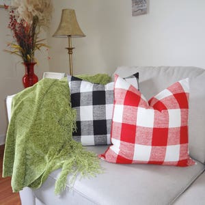 May include: Two throw pillows with a buffalo check pattern. One pillow is black and white, and the other is red and white. The pillows are on a white sofa with a green throw blanket.