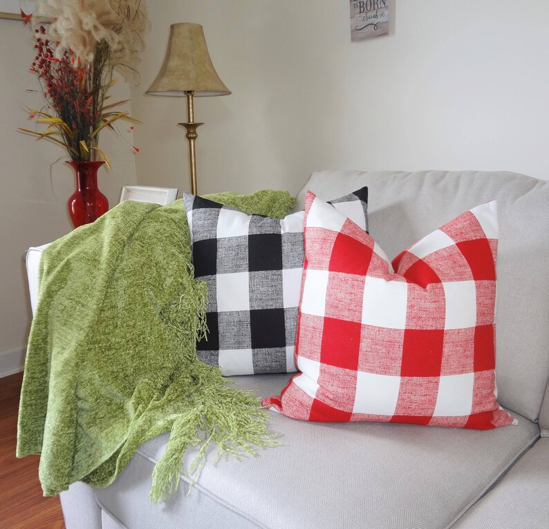 May include: Two throw pillows with a buffalo check pattern. One pillow is black and white, and the other is red and white. The pillows are on a white sofa with a green throw blanket.