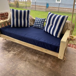 May include: A wooden porch swing with a blue cushion and two striped pillows. The swing is hanging from a rope and has a wooden frame.