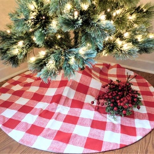 May include: Red and white buffalo check Christmas tree skirt. The skirt is round and has a white background with red squares. The skirt is placed on a wooden floor under a Christmas tree.