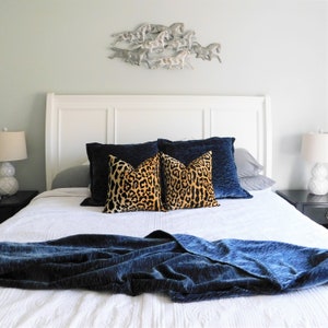 May include: A white bed with a blue velvet throw blanket and two leopard print throw pillows on top of a navy blue pillow. The bed has a white headboard and a white wall behind it. There is a silver metal sculpture of horses on the wall above the headboard.