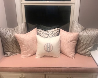 May include: A window seat cushion with a pink fabric cover and a variety of patterned throw pillows. The pillows are in shades of gray, pink, and white. The cushion is accented with a decorative trim.
