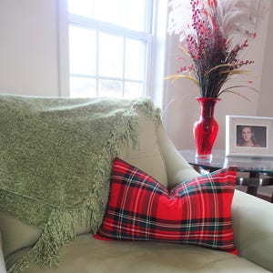 May include: A green throw blanket with fringe is draped over a light green armchair. A red and black tartan cushion is on the chair. A vase with red and white flowers is on a table next to the chair.