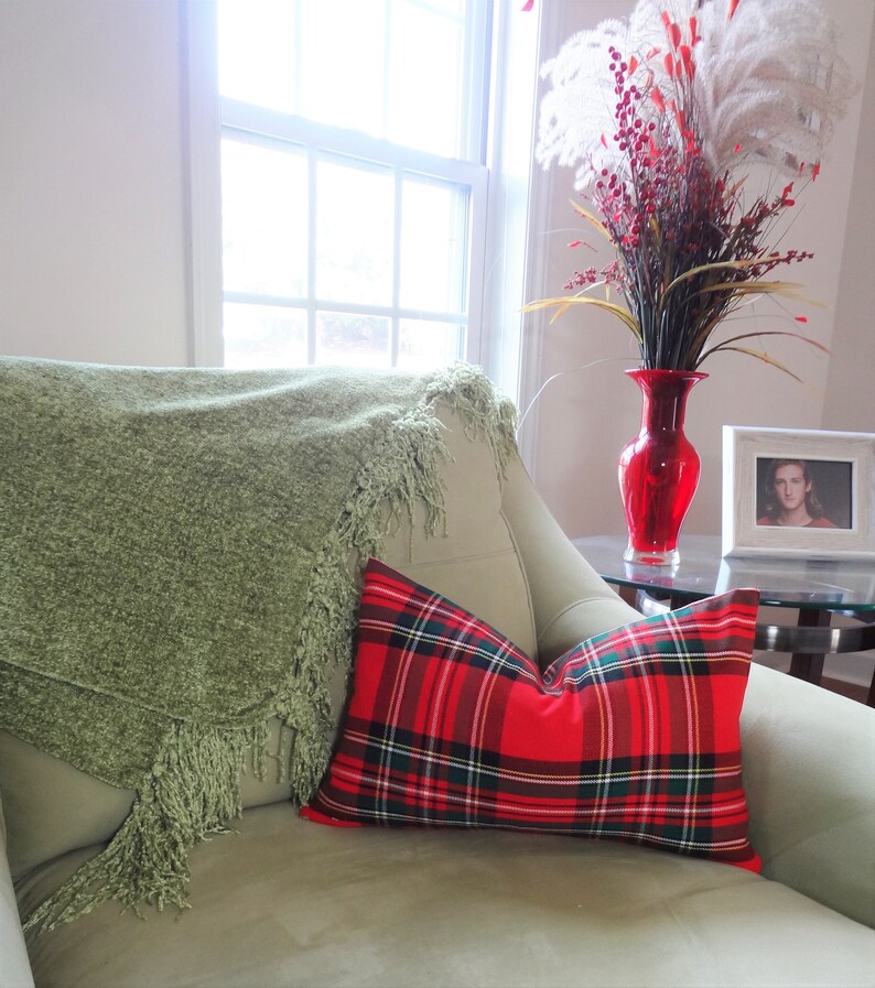 May include: A green throw blanket with fringe is draped over a light green armchair. A red and black tartan cushion is on the chair. A vase with red and white flowers is on a table next to the chair.