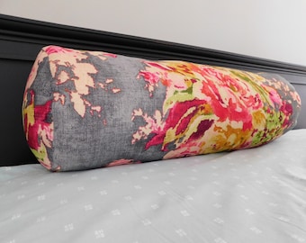 May include: A gray and white bolster pillow with a floral pattern in shades of pink, yellow, and green. The pillow is on a white bed with a subtle pattern.