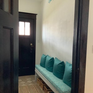 Puede incluir: Un banco blanco con un cojín turquesa y cuatro almohadas turquesa se encuentra en un pasillo con una puerta negra y una pared blanca. El banco tiene un estante debajo para almacenamiento.