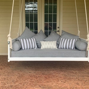 May include: White hanging porch swing bed with gray cushions and pillows. The swing is hanging from a white ceiling and is in front of a window with a view of trees.