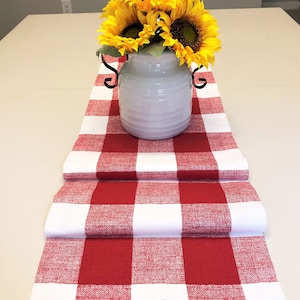 May include: Red and white checkered table runner with a white ceramic vase filled with yellow sunflowers.