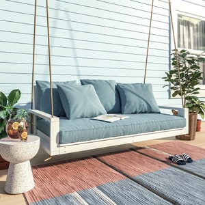 May include: White wooden swing bed with blue cushions and a book on the seat. The swing bed is hanging from the ceiling of a porch with a light blue wood siding. A small white side table with a glass pitcher of lemonade is in the foreground.